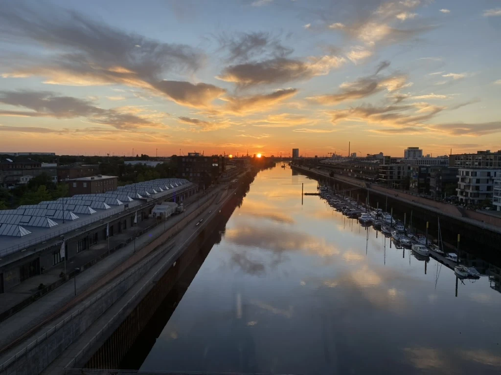 Sonnenuntergang über dem Bremer Hafen, Wasser spiegelt orange-rosa Himmel, Boote am Steg, Industriehallen am Ufer